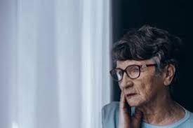 An elderly woman wearing glasses is looking out of a window.