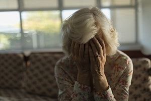 An elderly woman is sitting on a couch covering her face with her hands.