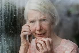 An elderly woman is talking on a cell phone while looking out of a window.