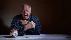 An elderly man is sitting at a table with an alarm clock.