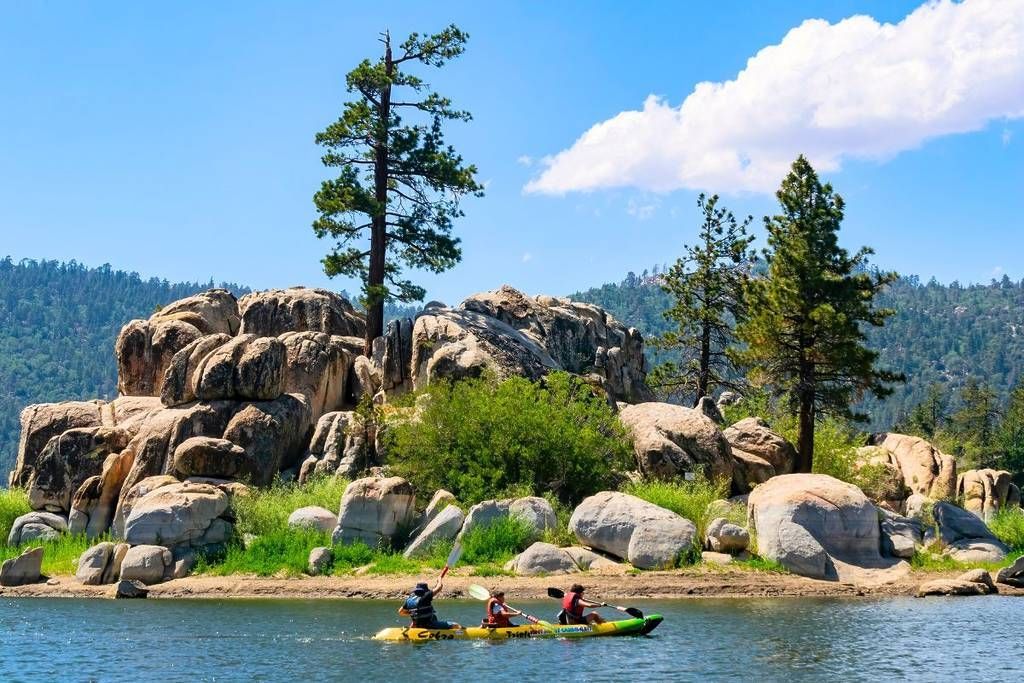 Kayakers paddle toward rocky shoreline with trees and blue sky in Big Bear Lake