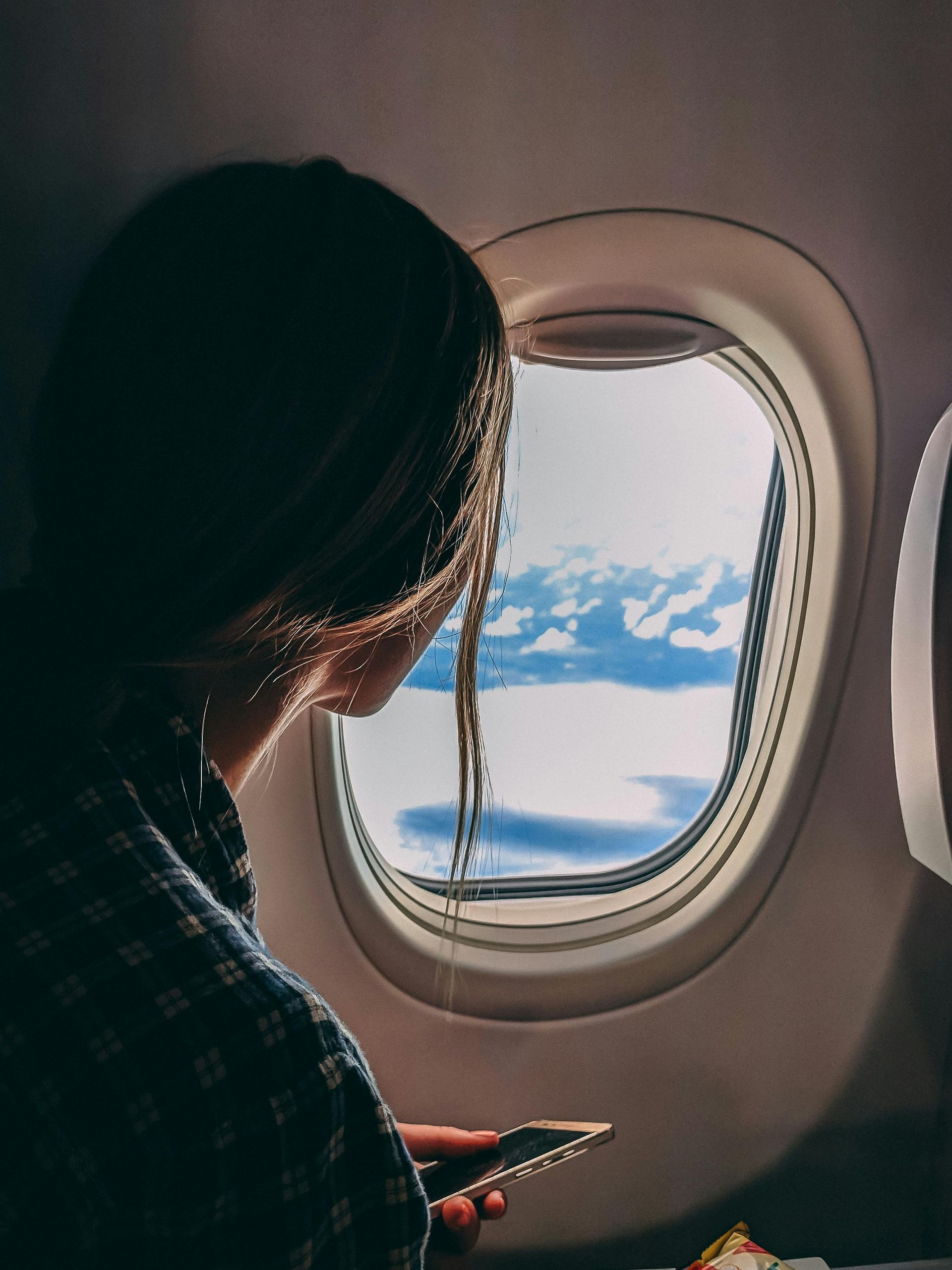 Person gazing out airplane window at clouds, holding phone.