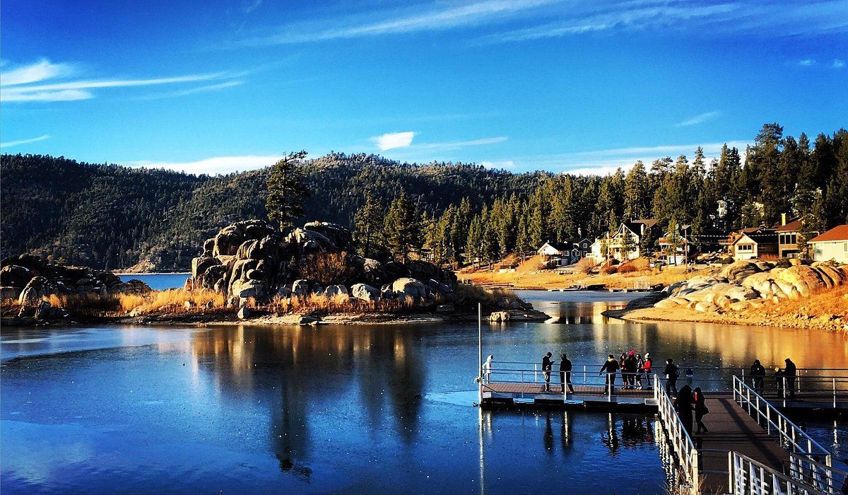 Big Bear Lake view with a wooden dock, people, rocks, trees, and homes under a blue sky.