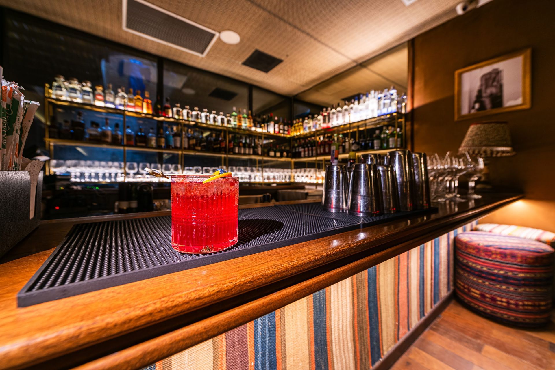 Bar com iluminação suave, garrafas coloridas, uma bebida vermelha no balcão e assentos listrados.