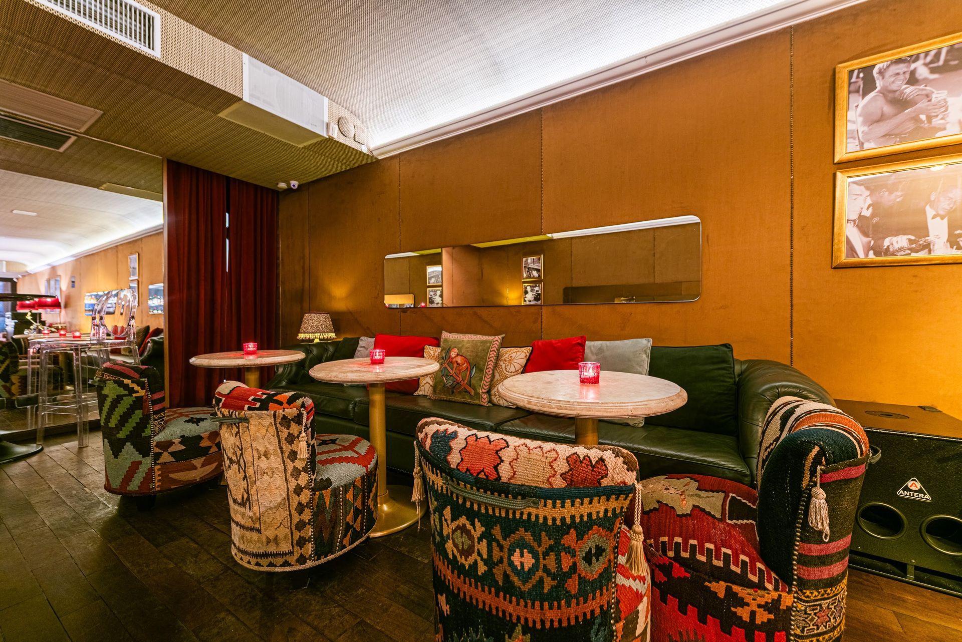 Sala de estar aconchegante com cadeiras estampadas, mesas redondas e iluminação laranja quente no interior de um restaurante.