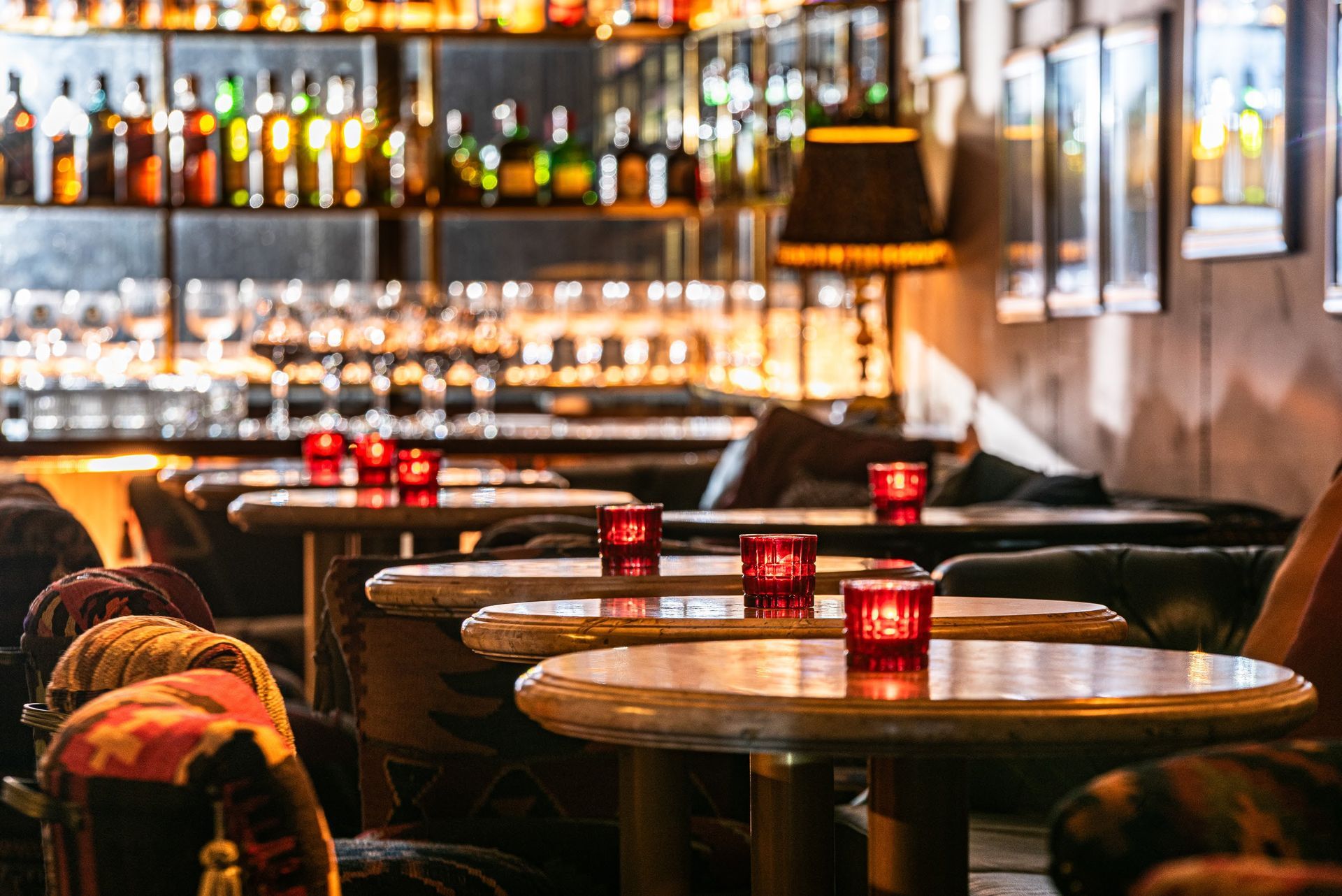 Interior de bar com iluminação suave, mesas, velas vermelhas e prateleiras de garrafas atrás do balcão.
