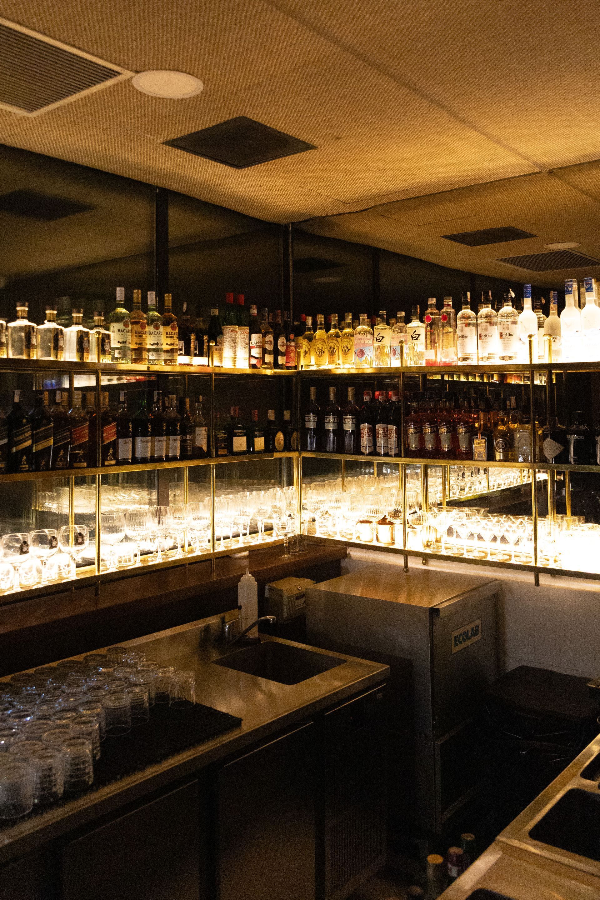 Interior de bar com iluminação suave, fileiras de garrafas em prateleiras iluminadas e um balcão em primeiro plano.