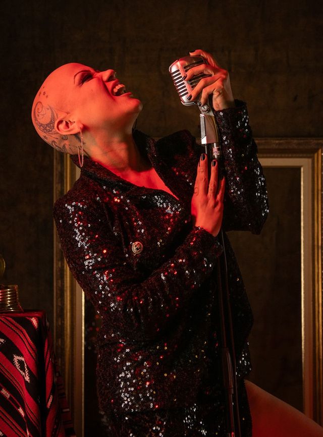 Cantora vestida com um traje preto de lantejoulas cantando em um microfone vintage sob iluminação vermelha de palco.