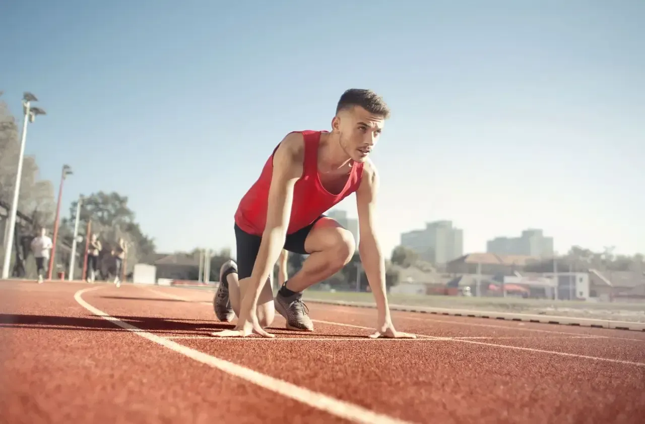 Runner in a red shirt crouches at the starting line on a track, preparing to sprint.