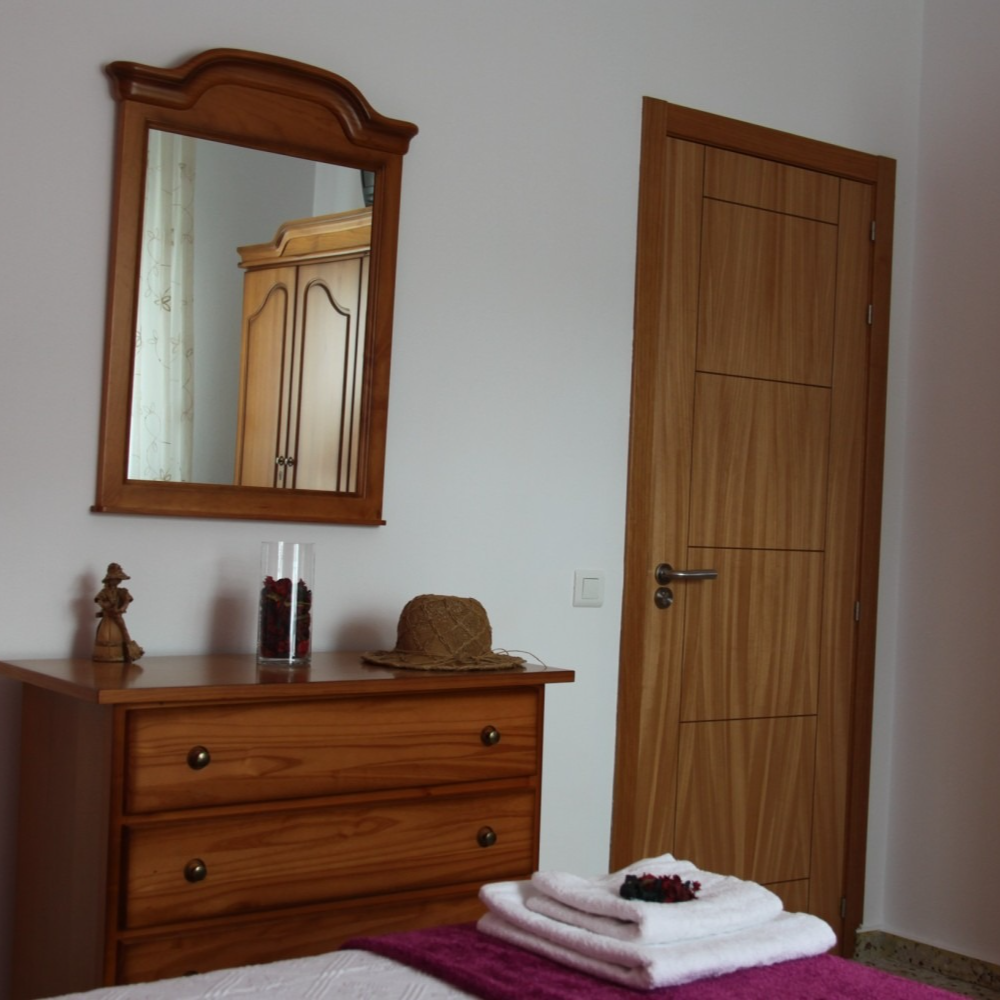 Wooden dresser with mirror and door in a bedroom, white walls, and a folded towel on the bed.