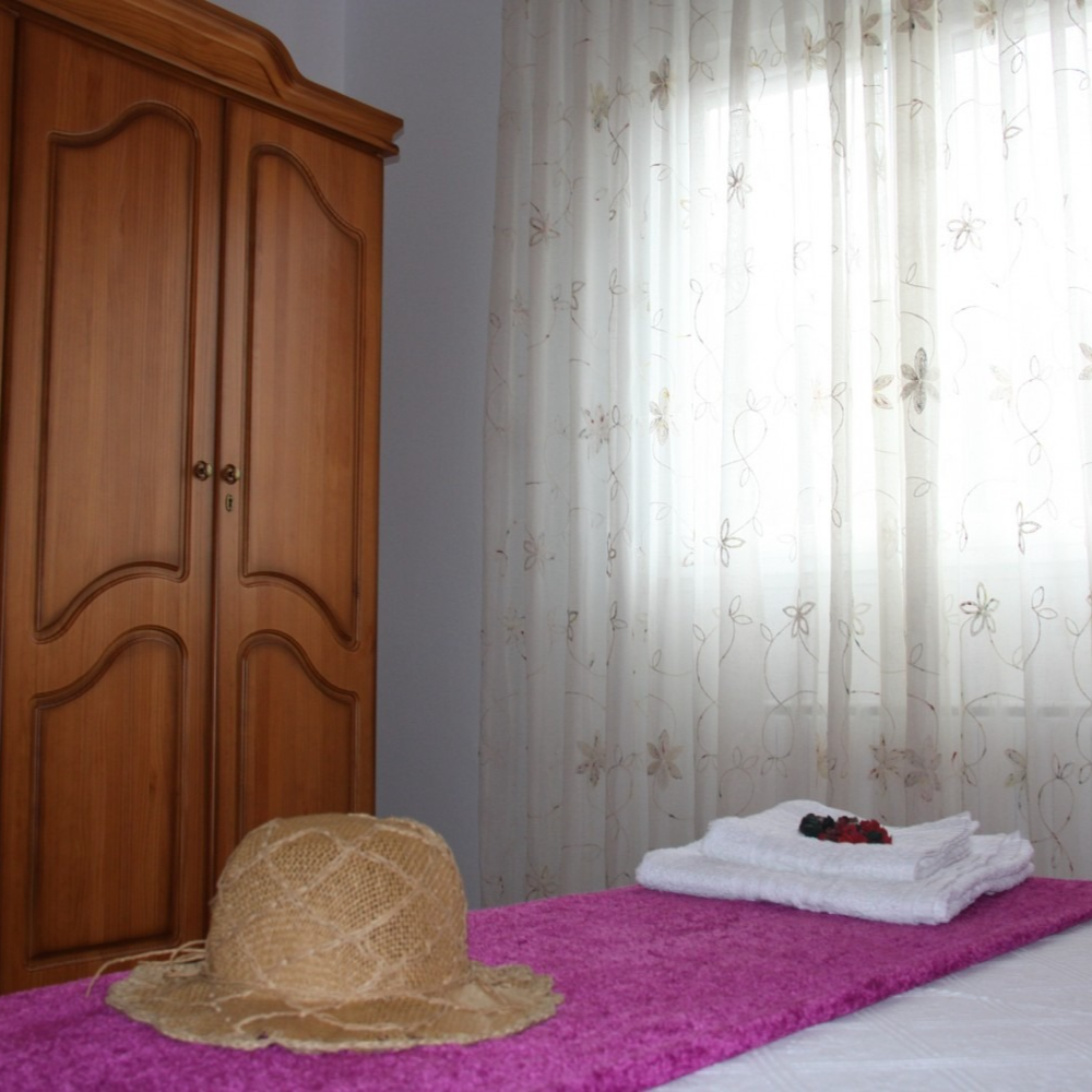 A bedroom with a wooden wardrobe, bed with towels, a hat, and a window with sheer curtains.