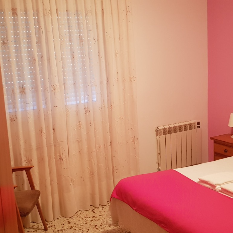 Bedroom interior with a window, bed, radiator, and chair. Pink wall and white curtains.