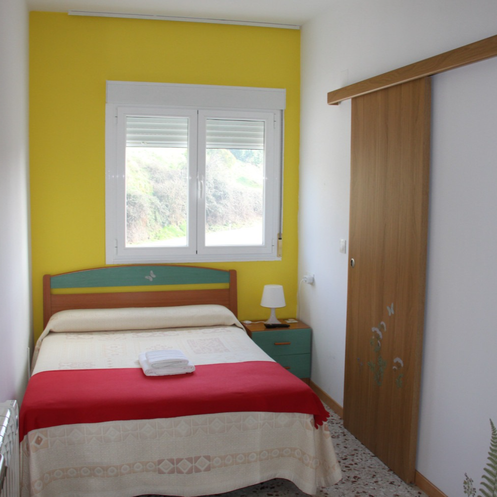 Bedroom with yellow wall, single bed with red blanket, small window, and wood closet door.