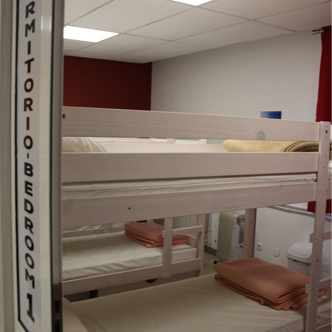 A hostel dormitory with three bunk beds, white walls, and a red accent wall; a sign reads 