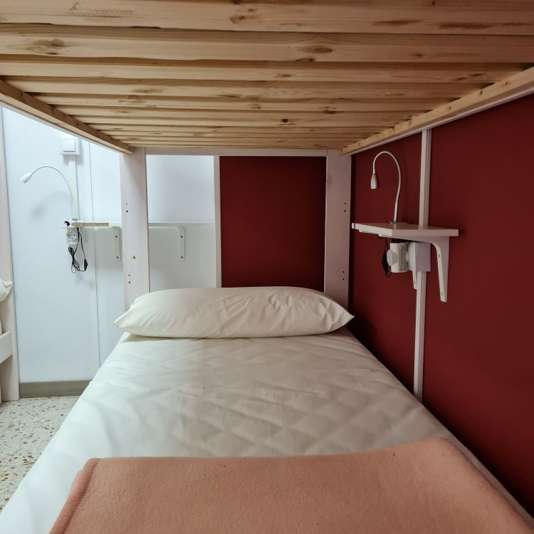 Single bed under a bunk bed frame. Red and white walls, reading lamps, and a peach-colored blanket.