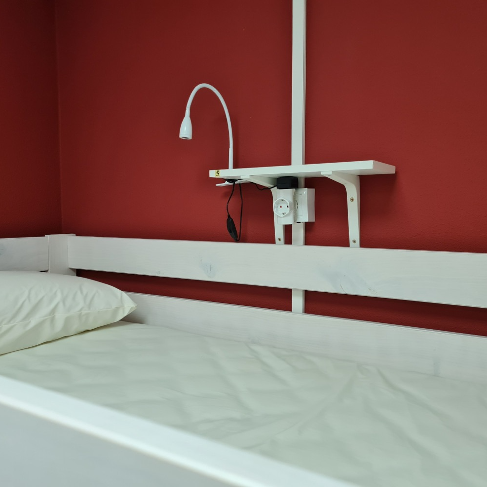 Bed with pillow and white linens against a red wall with a built-in lamp and outlet.