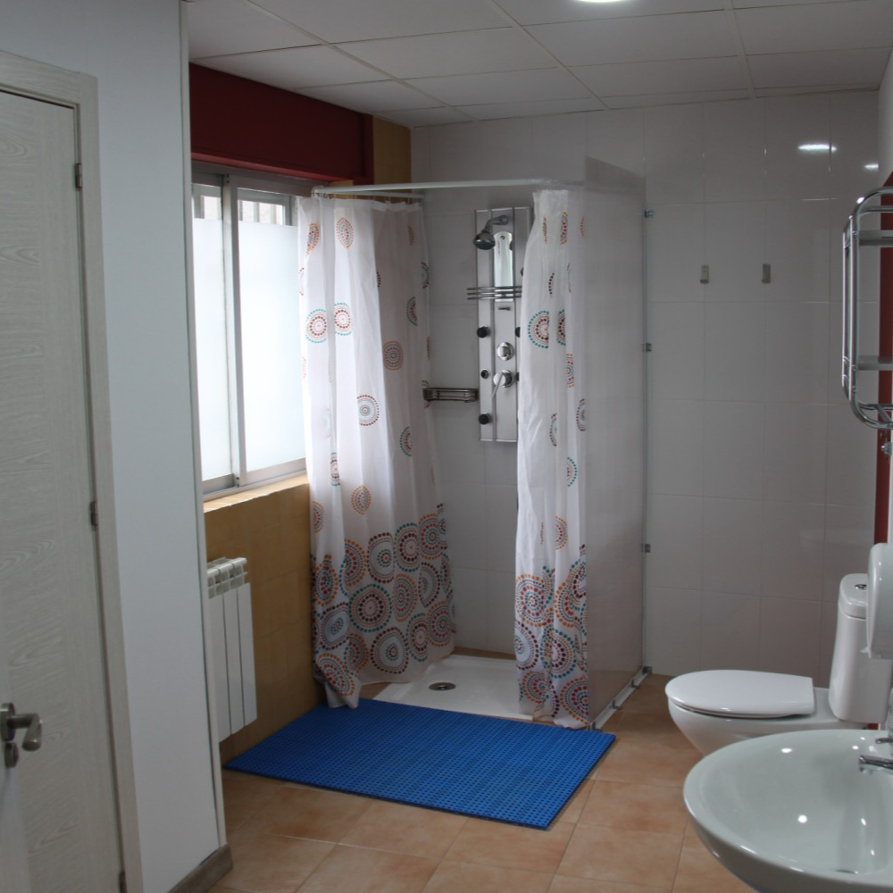 Bathroom with shower, toilet, sink, and window. Blue mat, white shower curtain, and tile floor.