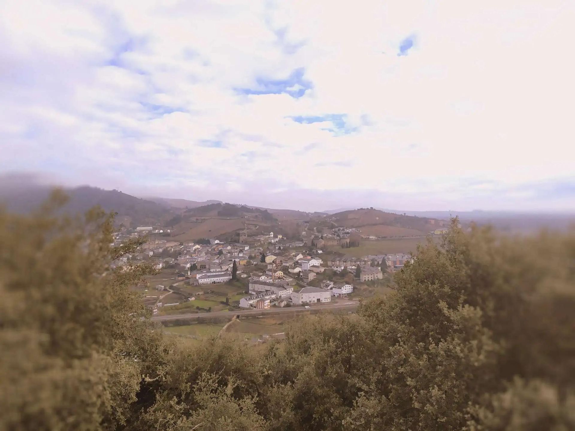 View of a town nestled in a valley, under a cloudy sky. Hills in the background, trees in the foreground.