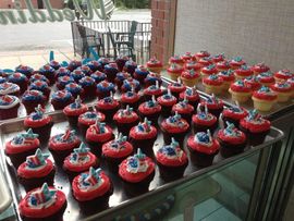 Rows of chocolate cupcakes with red frosting and blue toppers displayed on a glass table outdoors.