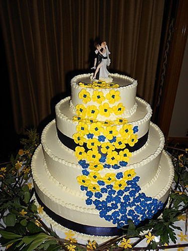 Four-tier white wedding cake with yellow and blue flower cascade and a bride topper