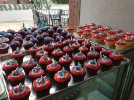 Rows of chocolate cupcakes with red frosting and blue toppers displayed on a glass table outdoors.