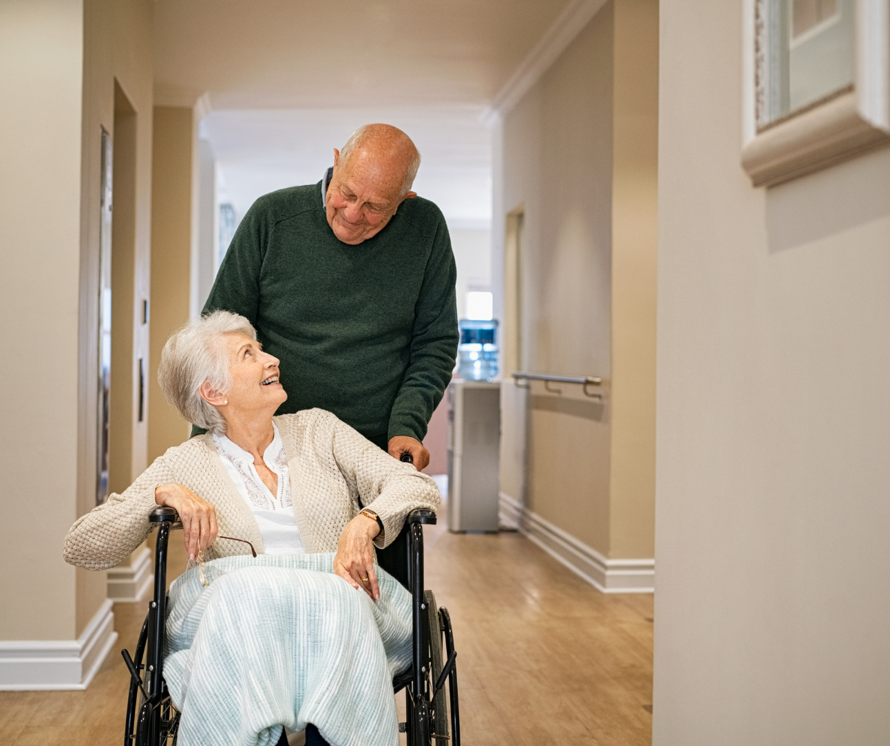 Elderly woman in wheelchair smiles at man pushing her in hallway.