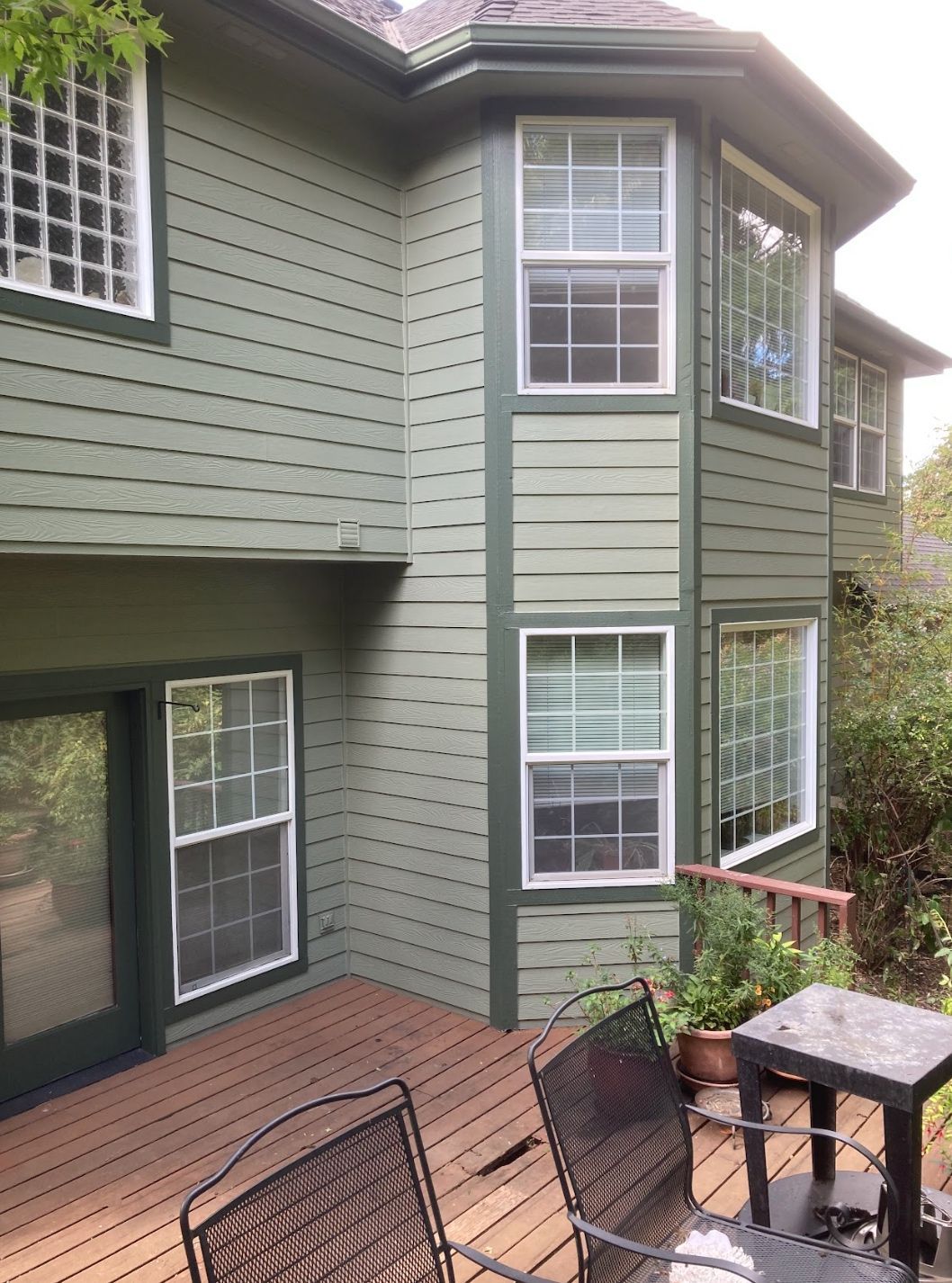 A two-story house with sage green horizontal siding, dark trim, and a wooden deck featuring outdoor furniture.