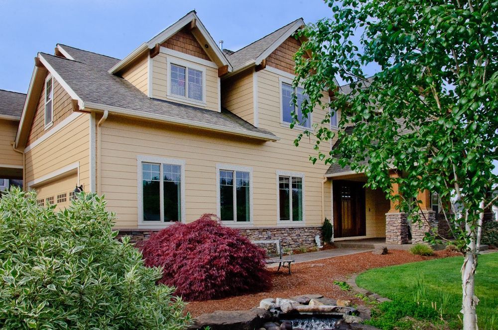 A two-story, beige suburban house with a brown shingled roof, landscaping, a red bush, and a light-colored tree out front.