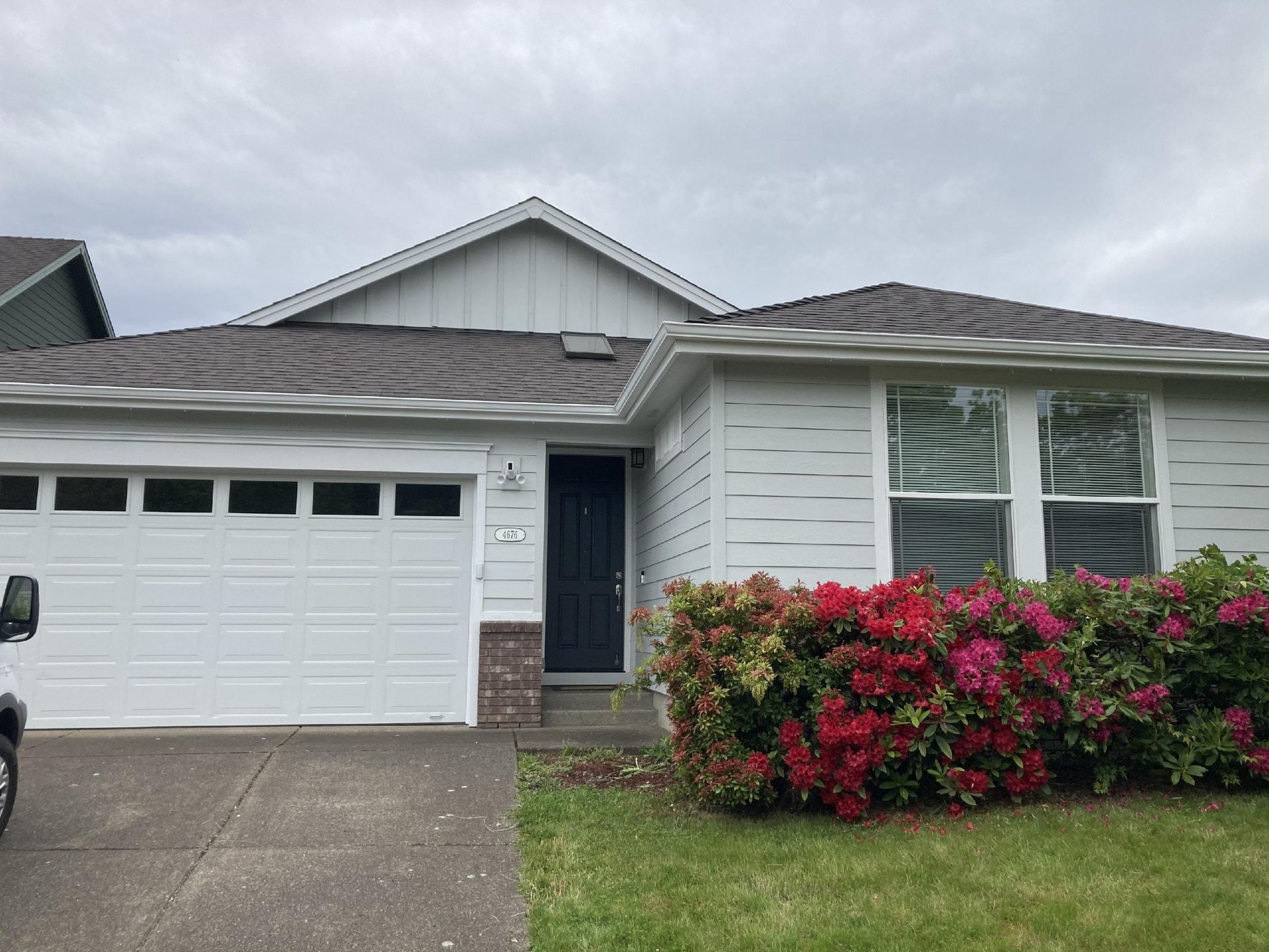 A light gray one-story house with a white garage door, dark front door, and a vibrant red azalea bush in the front yard.