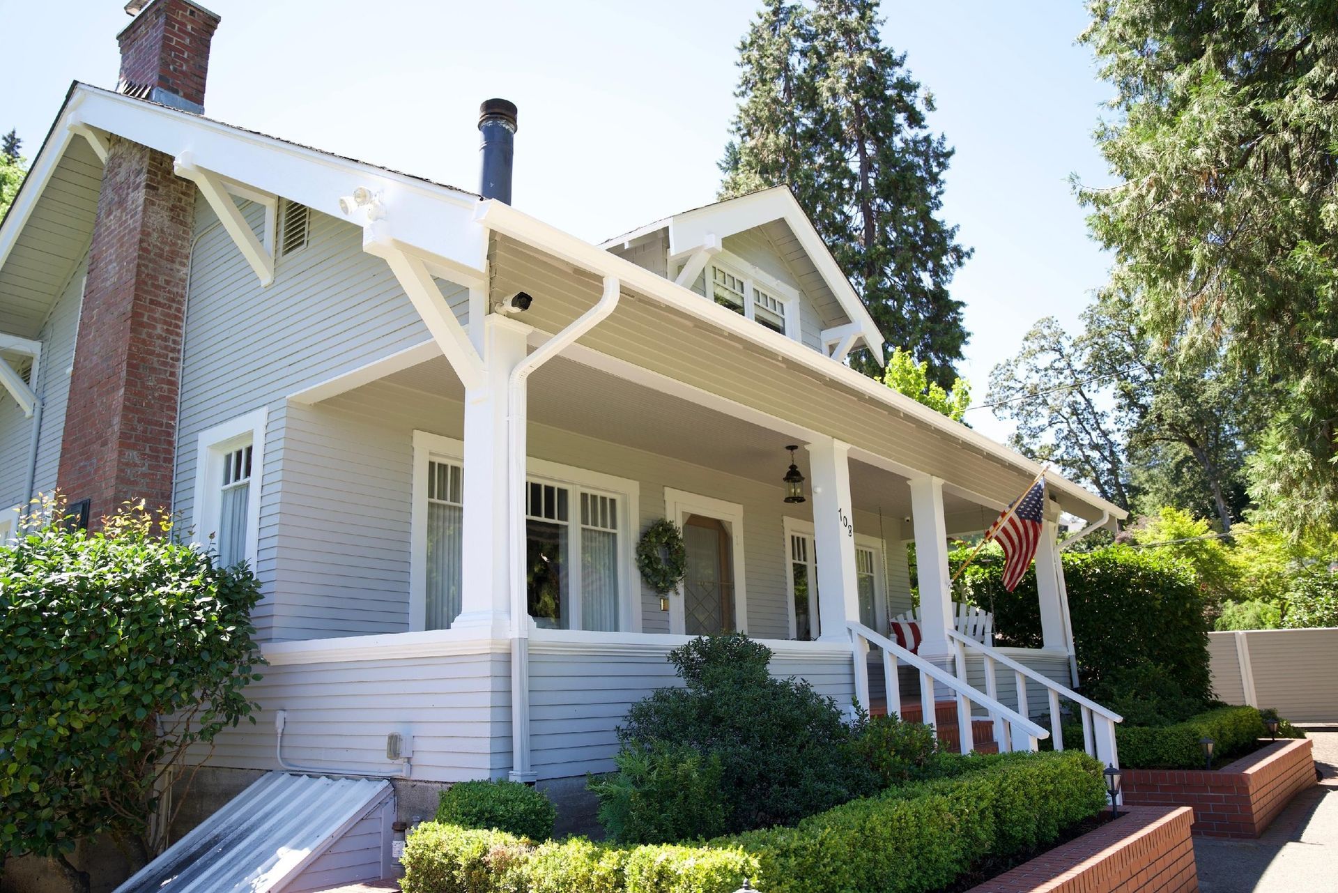 A light blue, one-story Craftsman-style house with a brick chimney, covered porch, white trim, and landscaped yard.