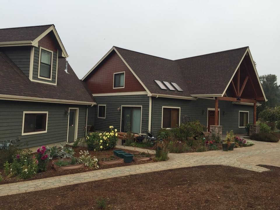 A two-story gray and reddish-brown house with a paved front walkway and a garden of blooming flowers.