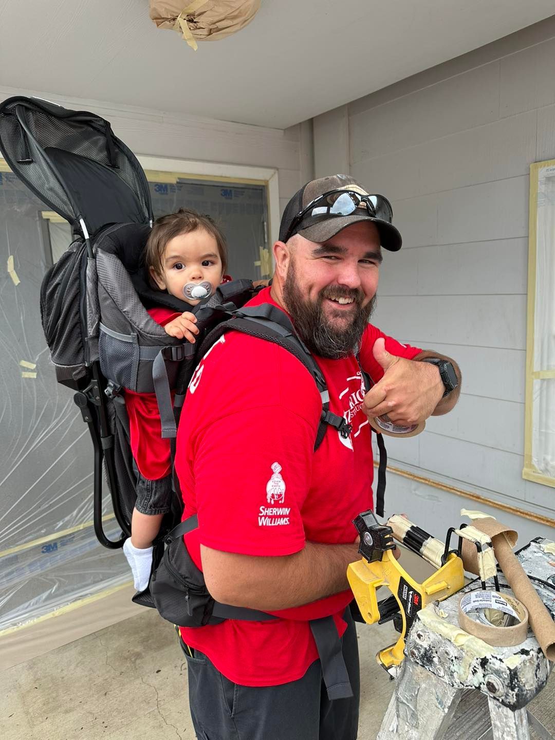 Man in red shirt carries a child in a backpack carrier, giving a thumbs up while painting.