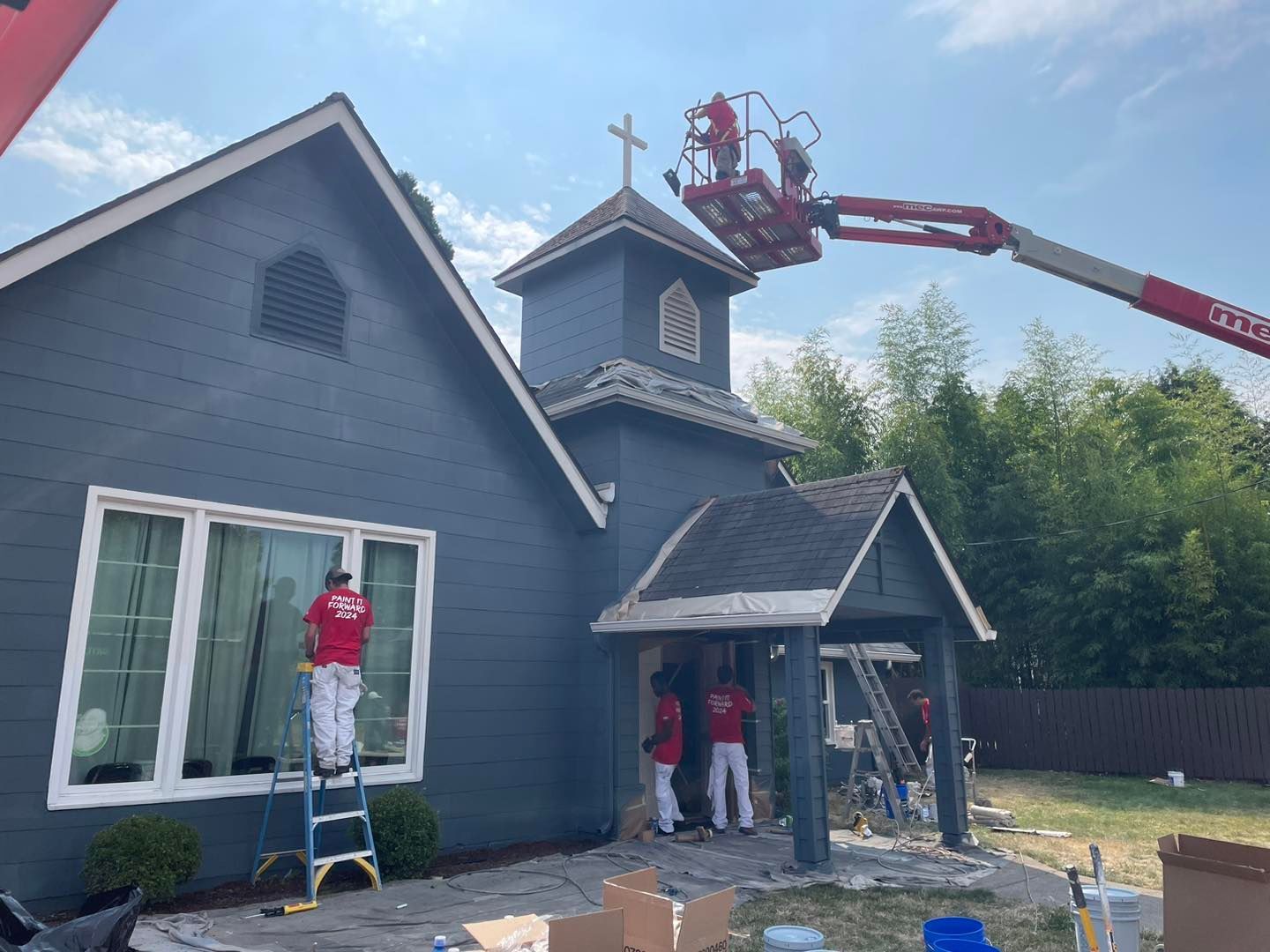 Exterior of a dark blue building being painted by workers using a lift and ladders on a sunny day.