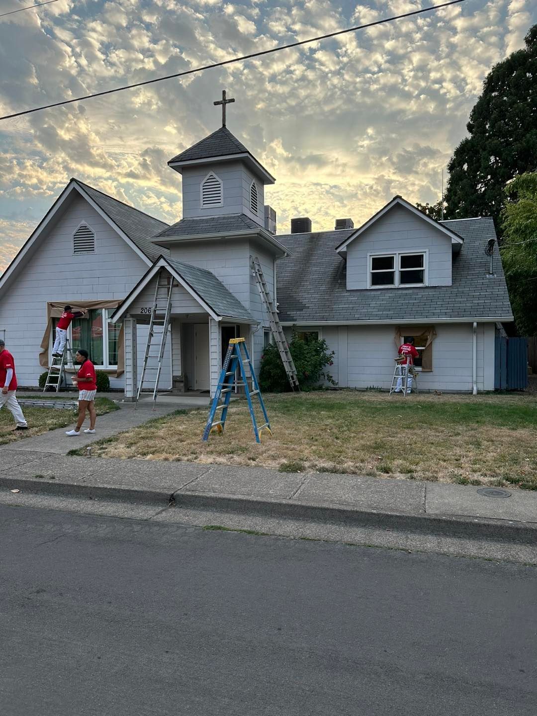Church building being painted by people on ladders; cloudy sky.