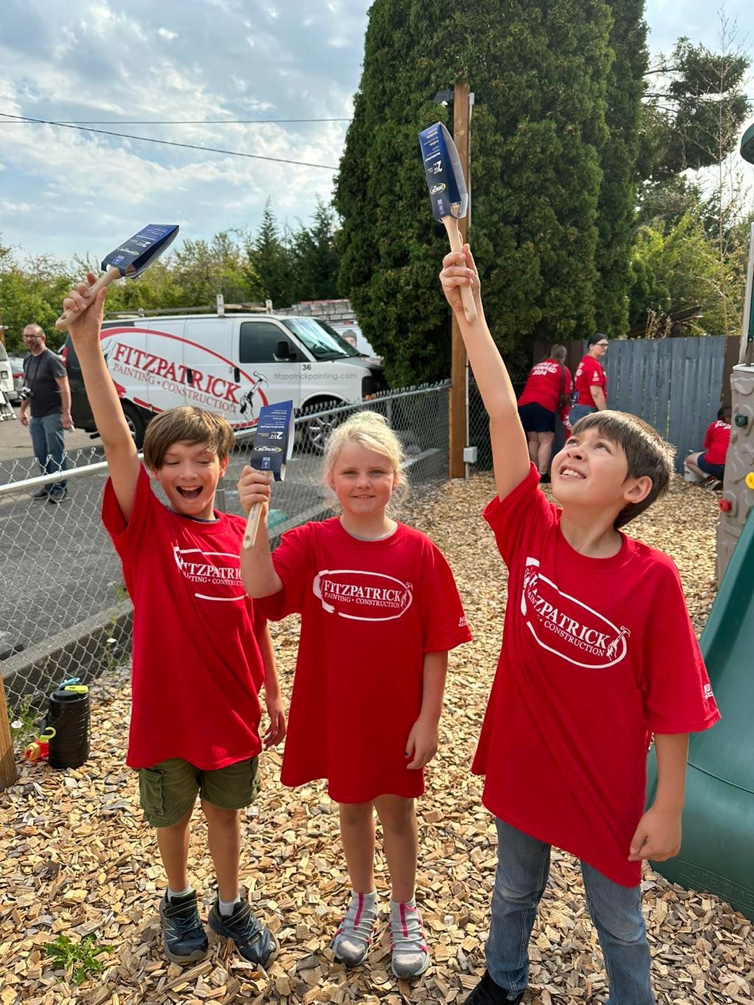 Three children in red shirts holding up paintbrushes outdoors.