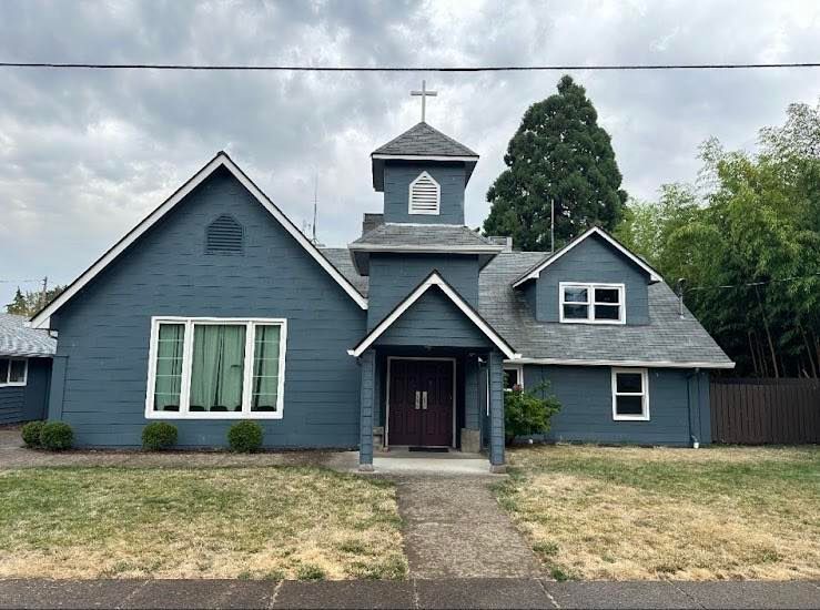 Blue church building with a cross on top, cloudy sky overhead, and a walkway leading to the entrance.