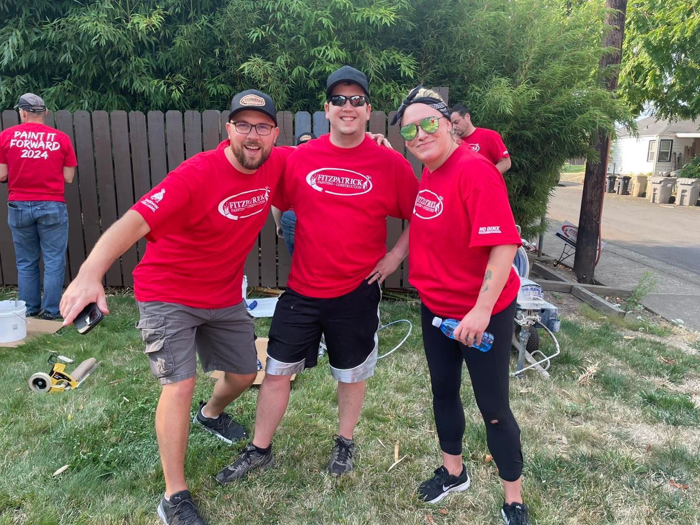 Three people in red shirts and hats pose outside, grass and fence in background, other people.