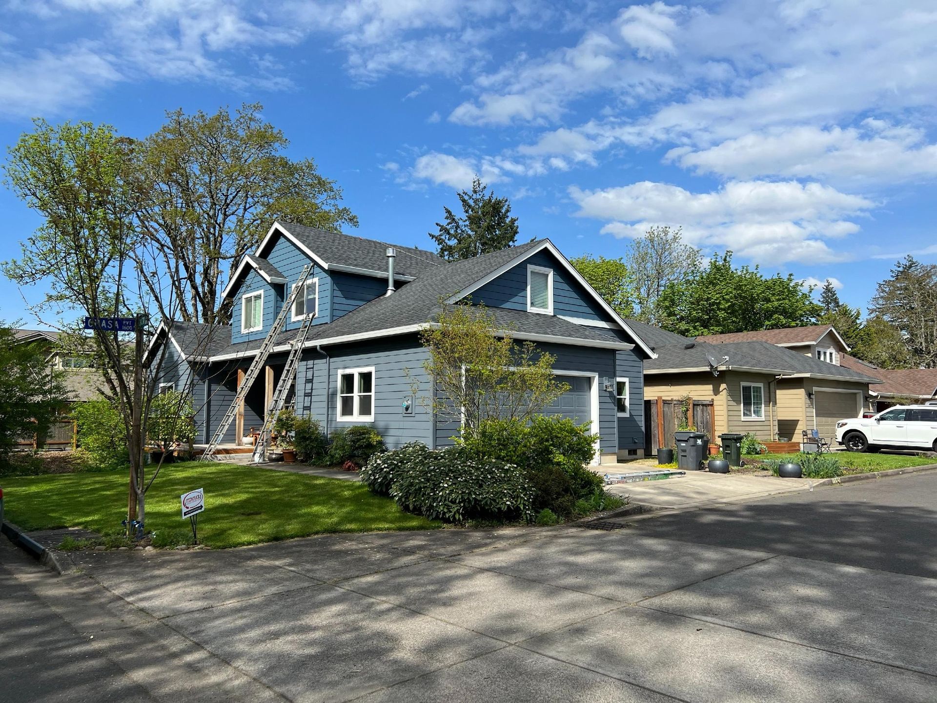 A blue, two-story house under construction with scaffolding against the exterior, surrounded by trees and a sunny sky.