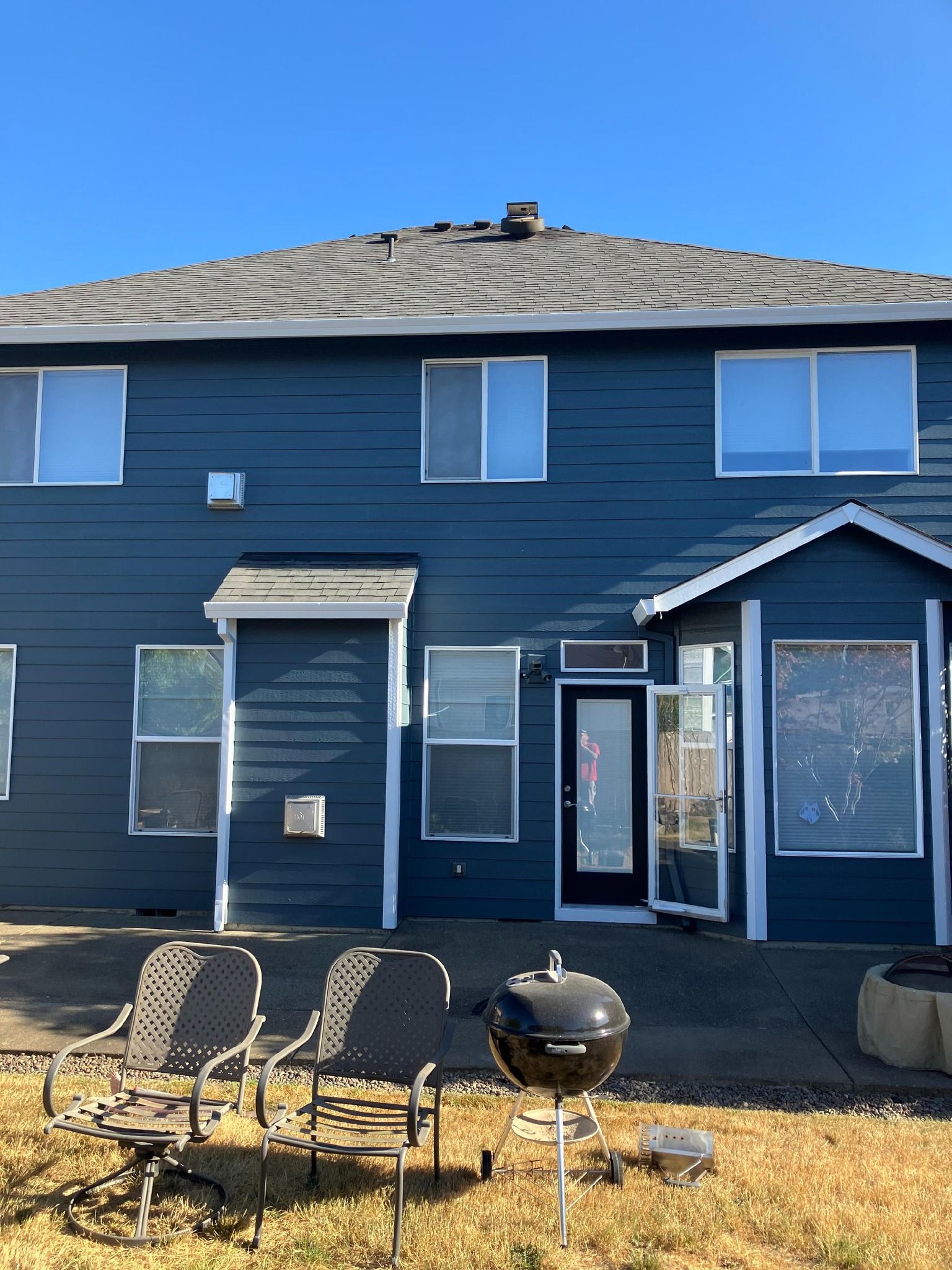 Back exterior view of a blue, two-story house with a patio, two chairs, and a charcoal grill on a dry, grassy yard.
