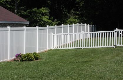 White vinyl fence with a gate along a green lawn beside a house and trees