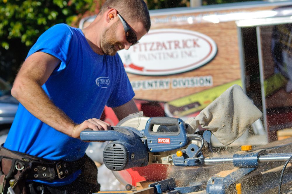 Carpenter using a blue miter saw to cut wood outdoors; Fitzpatrick Painting Inc. sign in the background.