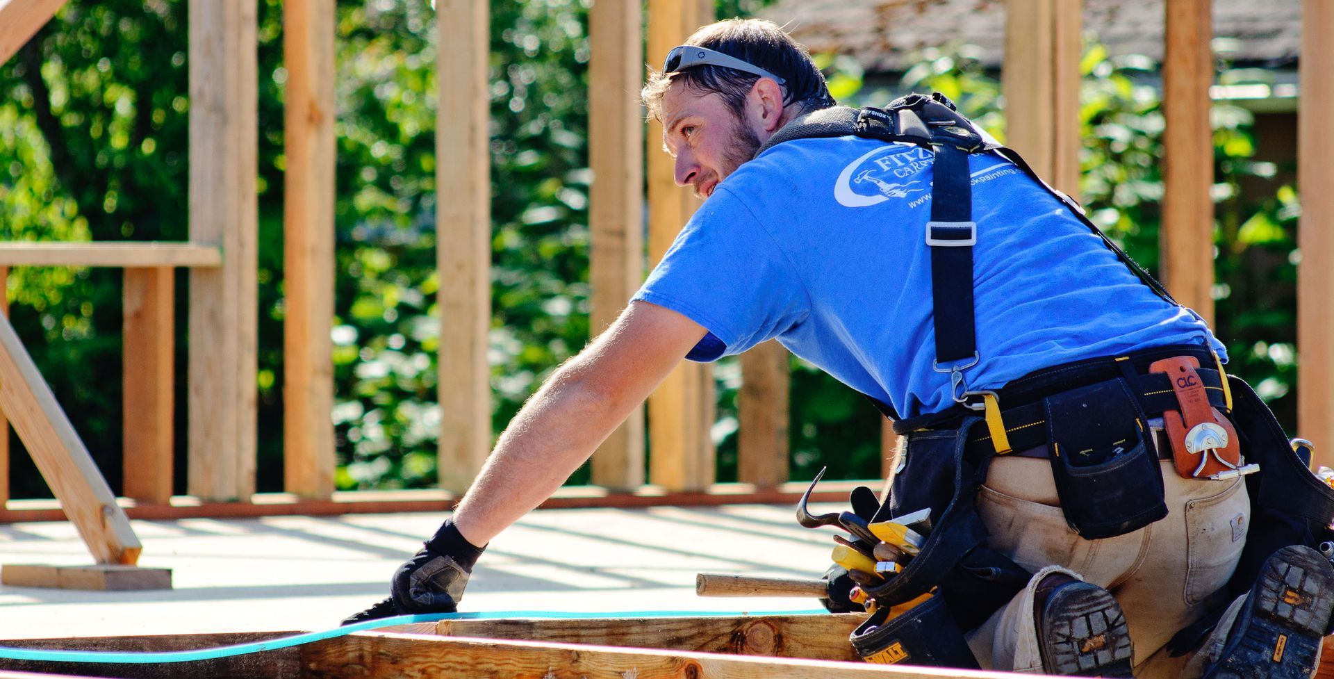 Construction worker in a blue shirt and tool belt, working on wooden framing outdoors.