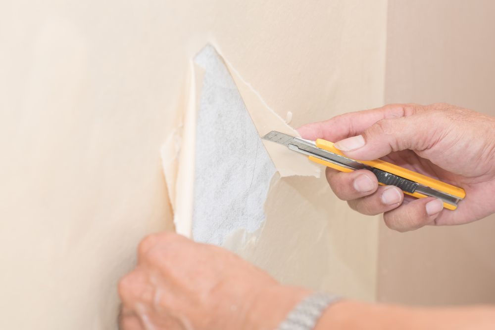 Hands peeling wallpaper, using a utility knife to cut away a section. Light tan wall.