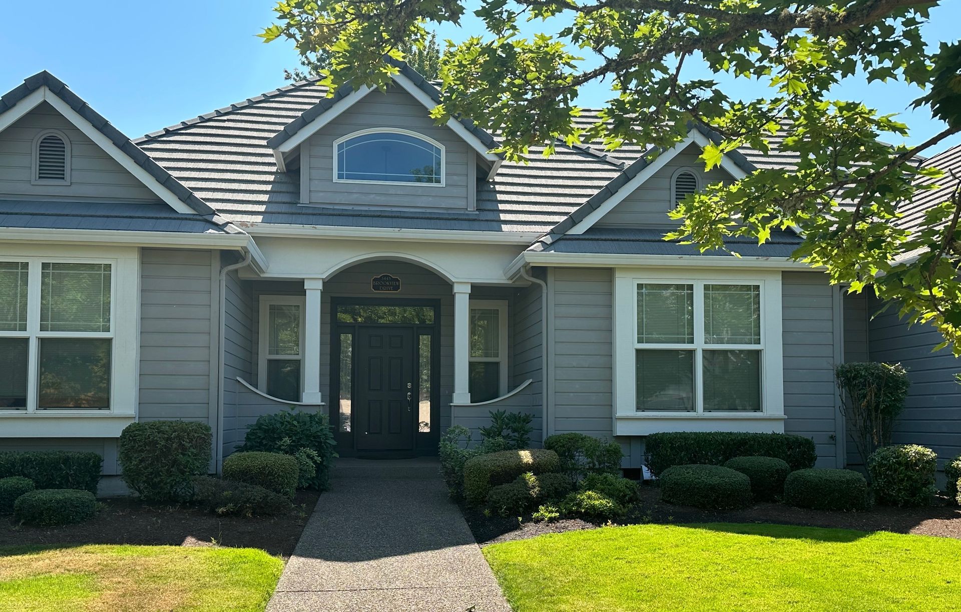 Gray house with a gray tile roof, dormer, and arched front entrance, with green bushes and grass.