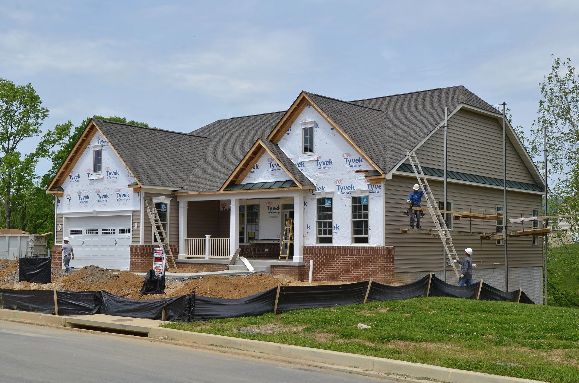 Construction workers build a house with brick base, siding, and gabled roof on a sunny day.