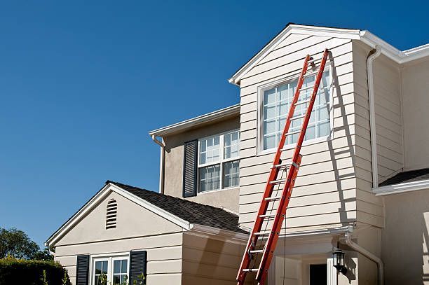 A bright orange extension ladder leaning against the beige siding of a two-story house under a clear blue sky.