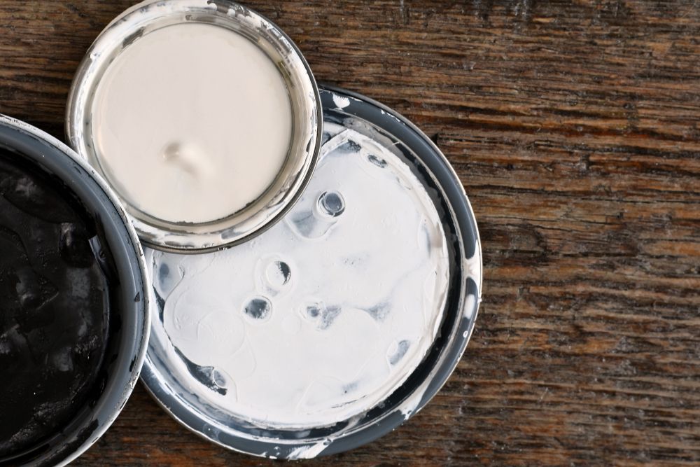 Black and white paint cans on a wooden surface; white paint on lid, drips on tray.