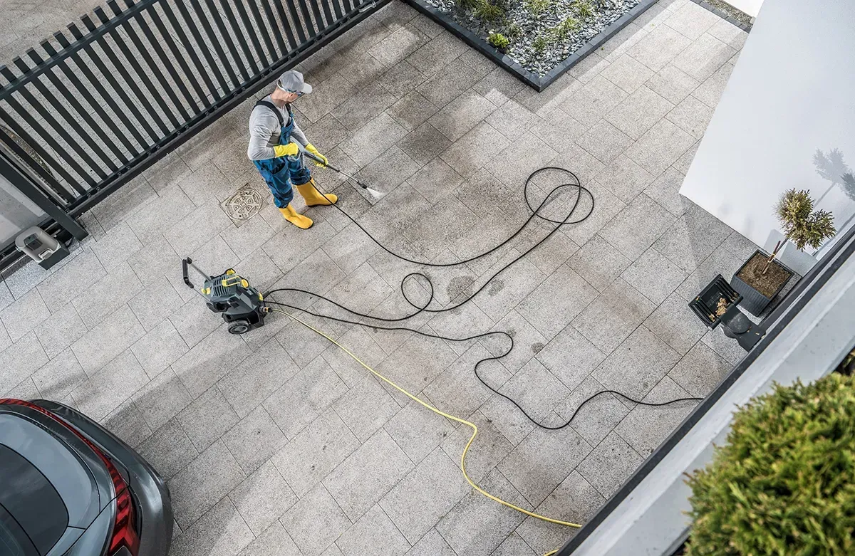 Worker pressure-washing a driveway beside a parked car and a gate, with hoses stretched across the pavement