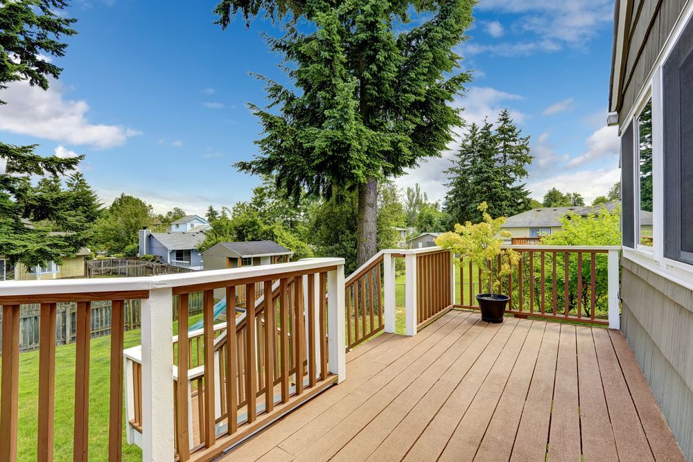 Wooden deck with brown railings, overlooking a green yard and houses, blue sky in background.