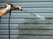 Hand holding a pressure washer, cleaning mold off of gray siding.
